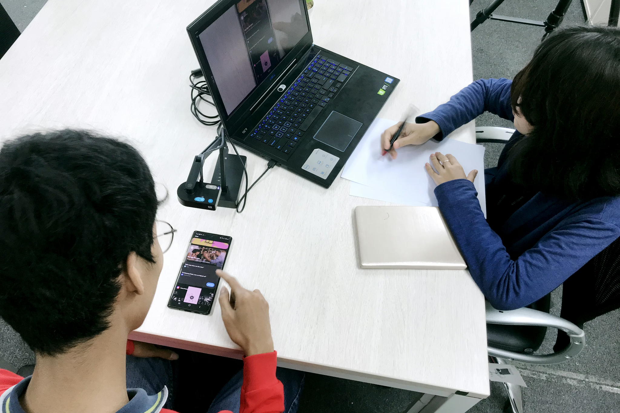 Overhead view of a moderated in-person usability test, where a participant uses a mobile app on a smartphone while being filmed by a small camera; the live feed appears on a nearby laptop as a moderator observes and takes notes at the desk.