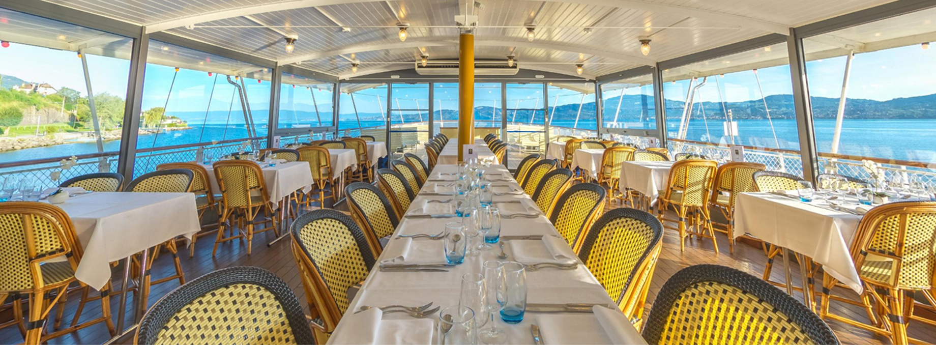 Interior dining area aboard a CGN ferry, featuring elegantly set tables and panoramic views of Lake Geneva through large windows.