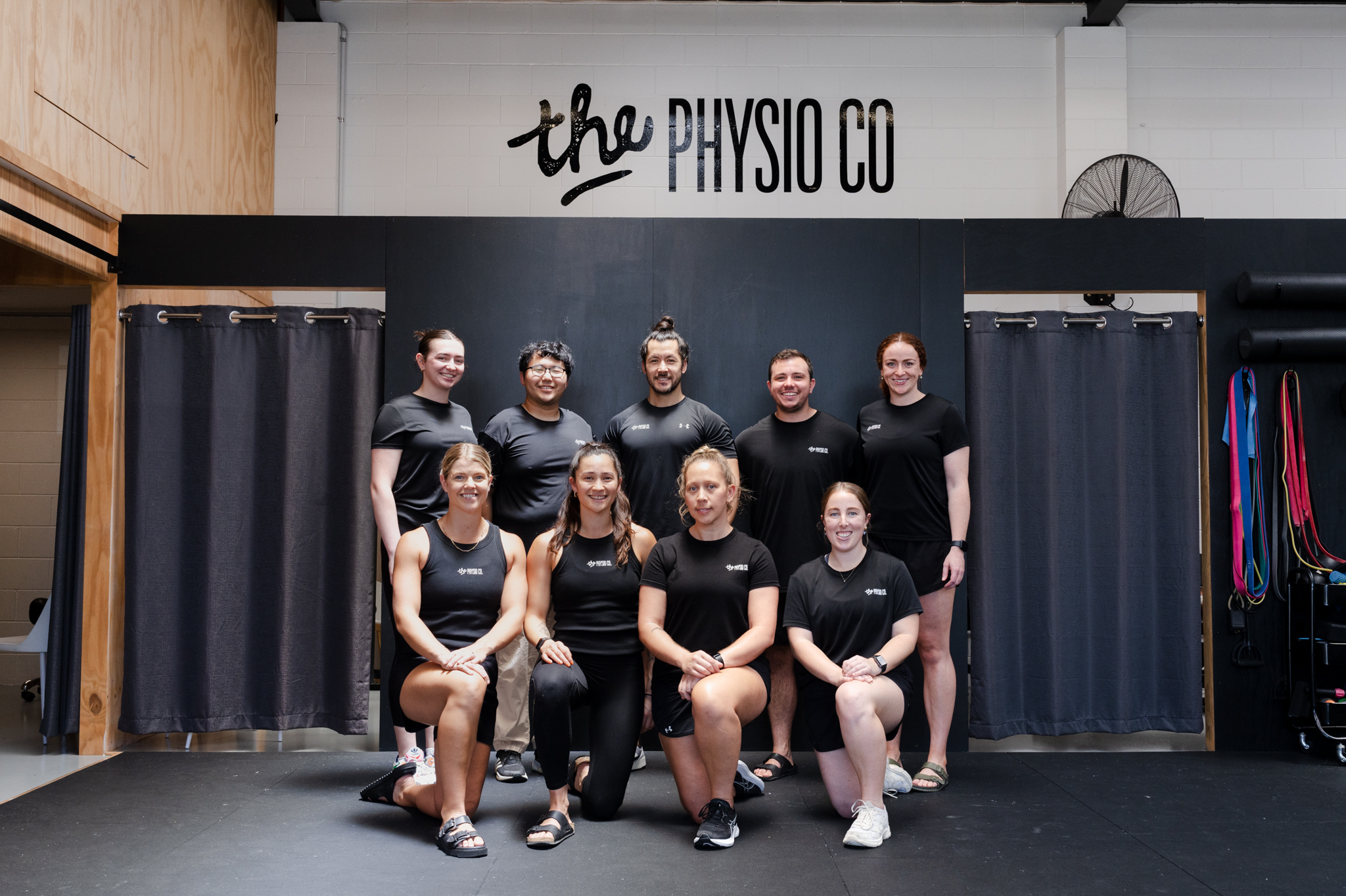 The Physio Co. team of physiotherapists and staff posing together in matching black t-shirts beneath the clinic's logo on the studio wall.