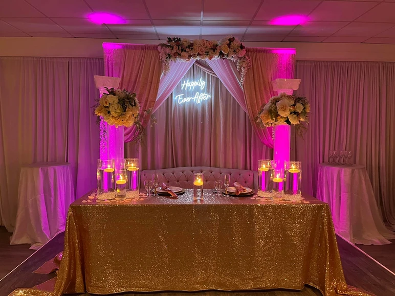 Romantic setup with a gold table cover, bride and groom chair, pink and white floral decorations, candles, a neon sign saying 'Happily Ever After,' and pink curtains in the background at The Palmyra Venue in New Jersey