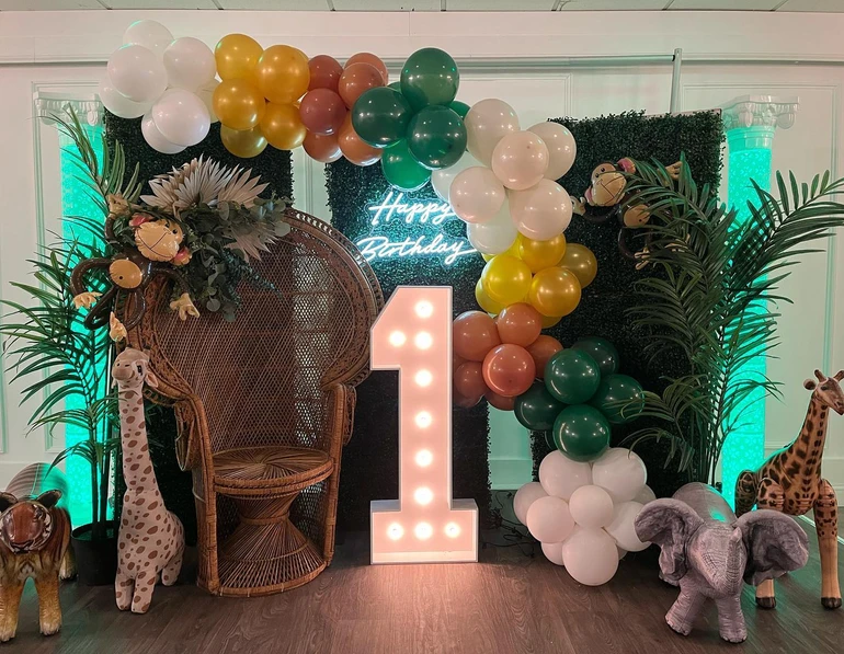 Safari-themed 1-year-old celebration featuring an illuminated number 1, next to brown chair. Grass mural and vibrant balloons at  The Palmyra Venue in New Jersey