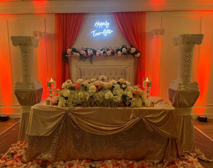 Elegant setup at The Palmyra Venue in New Jersey, featuring a gold table cover and chairs for the bride and groom. Adorned with white, orange, and pink floral decorations, a neon sign displaying 'Happily Ever After,' and complemented by vibrant orange curtains in the background