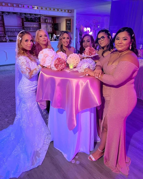 Gorgeous bride in her beautiful wedding dress accompanied by four women dressed in pink, standing at The Palmyra Venue next to a table adorned with varying shades of pink flowers and holding a pink and white bouquet
