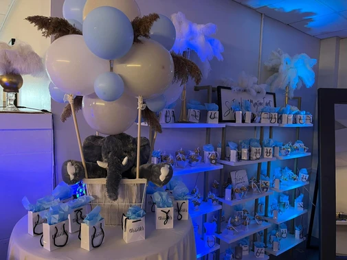 Wall with gift bags for guests next to a table adorned with an elephant stuffed animal decoration and balloons, complementing the light blue and gray themed decorations at The Palmyra Venue.