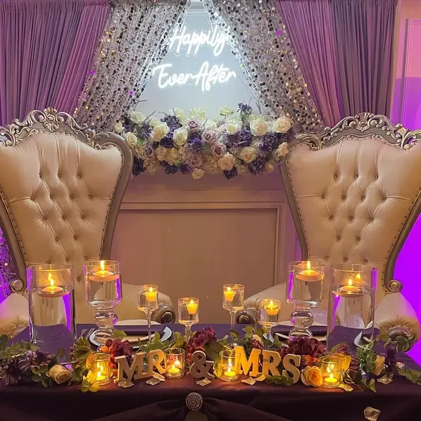Two exquisite beige chairs adorned with a stunning wedding floral arrangement, complemented by elegant purple curtains in the background at The Palmyra Venue in New Jersey. A captivating neon sign displaying 'Happily Ever After' adds a touch of enchantment.