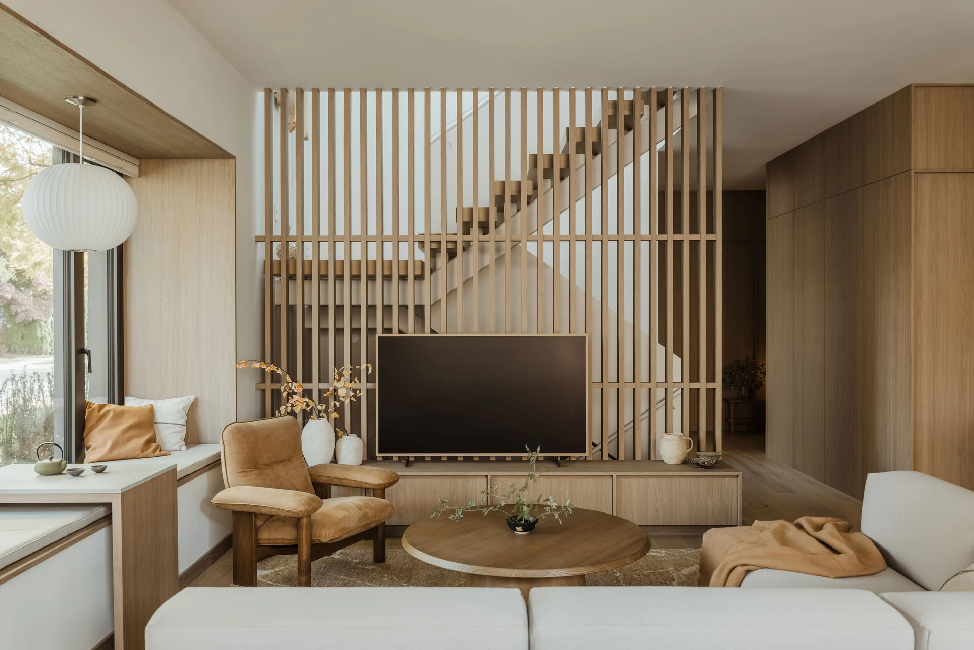 Modern living room with wooden slat wall, beige armchair, round wooden coffee table, and large flat-screen TV.