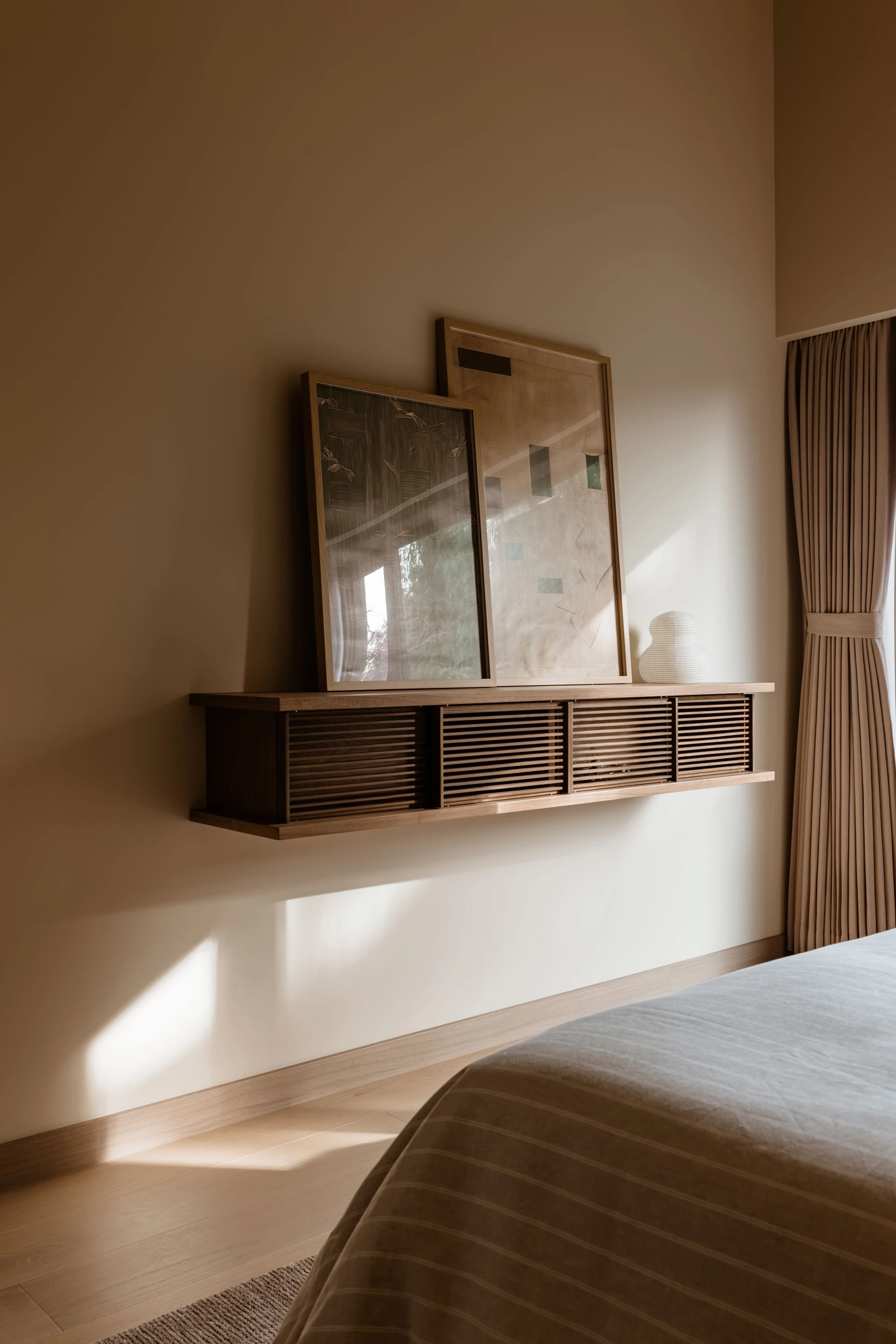 Minimalist bedroom corner with a wall-mounted wooden shelf holding two framed artworks and a decorative white vase, with beige curtains and a bed partially visible.