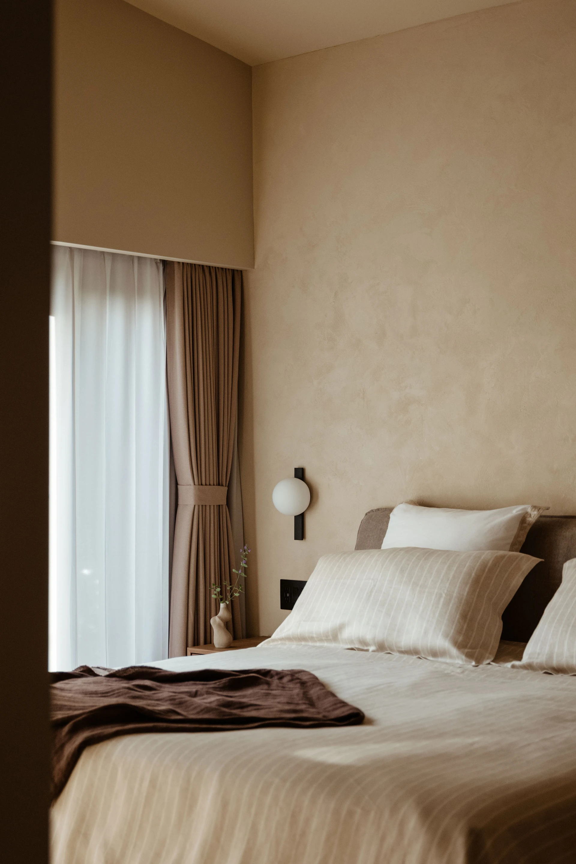 Cozy bedroom with beige walls, striped bedding, brown throw, and a small vase with flowers on a bedside table near a window with curtains.
