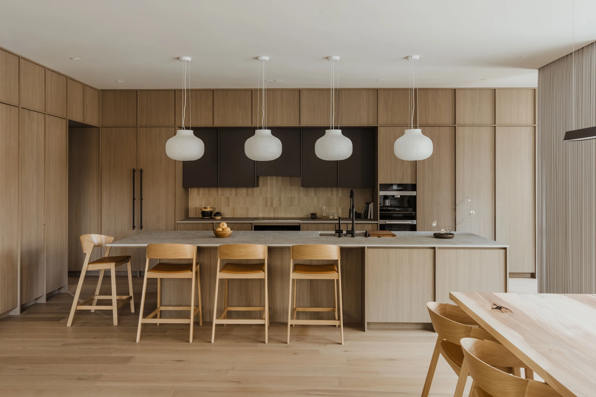 Modern kitchen with light wood cabinetry, a large island with four wooden chairs, stone countertop, four white pendant lights, and a wooden dining table with chairs.
