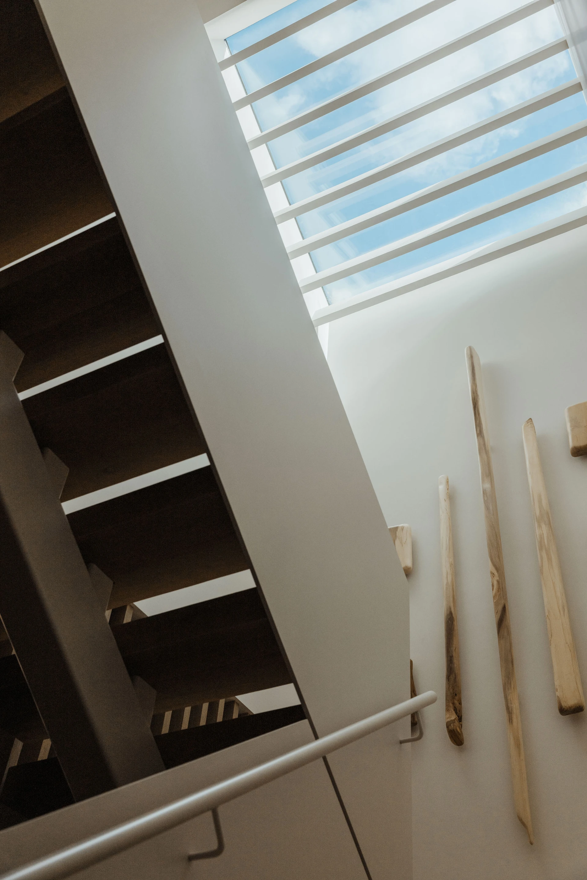 Modern indoor staircase with wooden steps, a white wall featuring vertical wooden sculptures, and a skylight with horizontal beams.