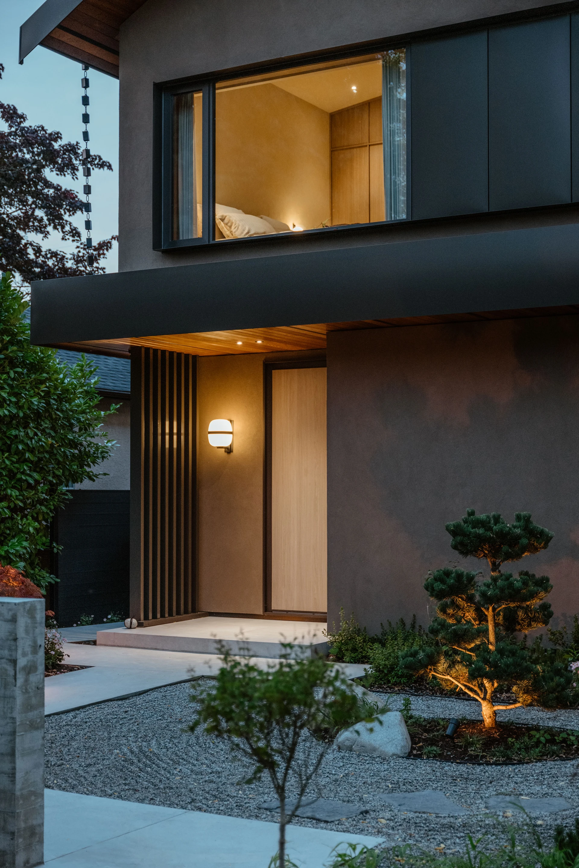 Modern house entrance at dusk with illuminated porch light, large window showing a bedroom, and landscaped garden with small trees and gravel pathway.