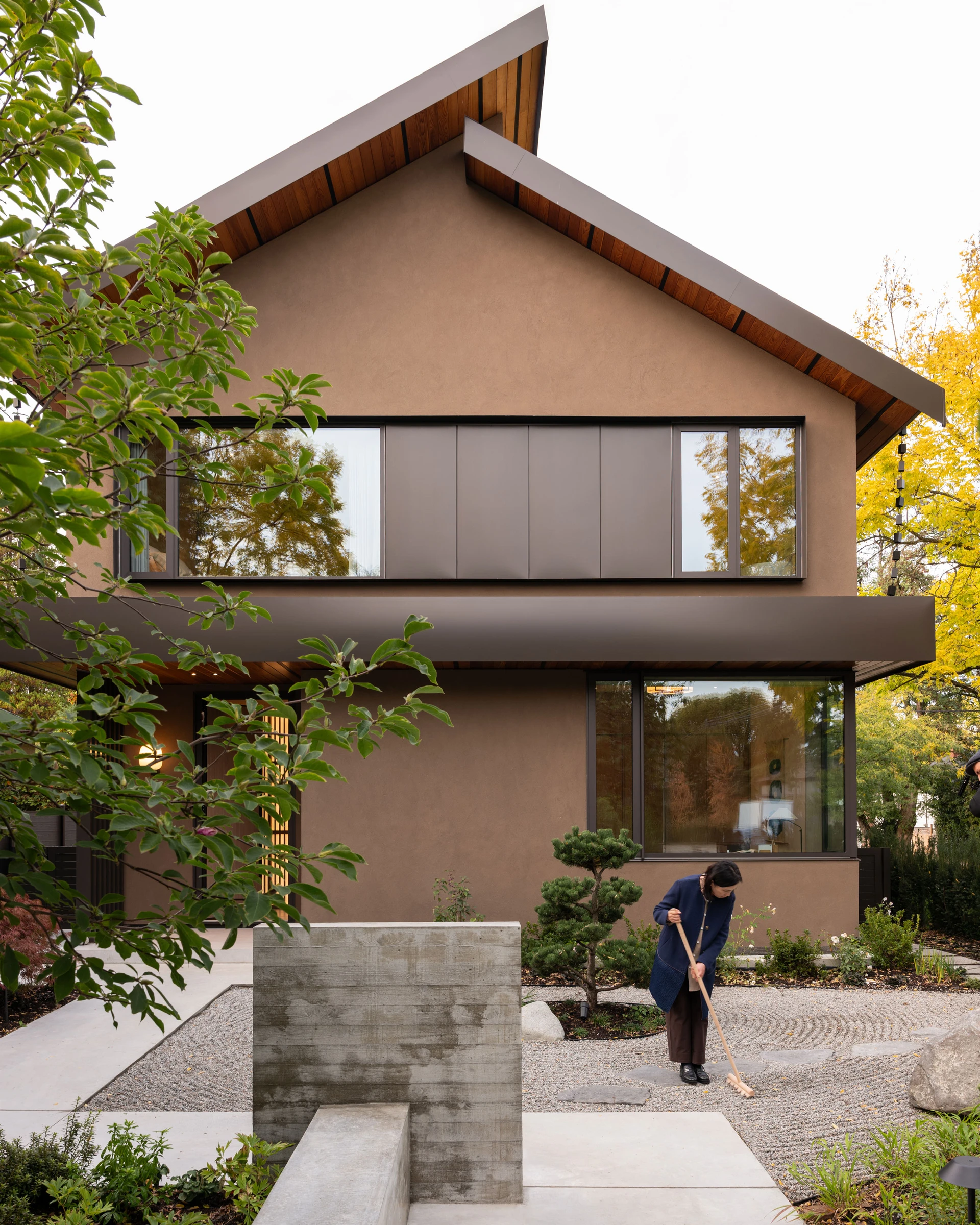 Modern two-story brown house with large windows and a person raking gravel in a landscaped front yard with greenery.