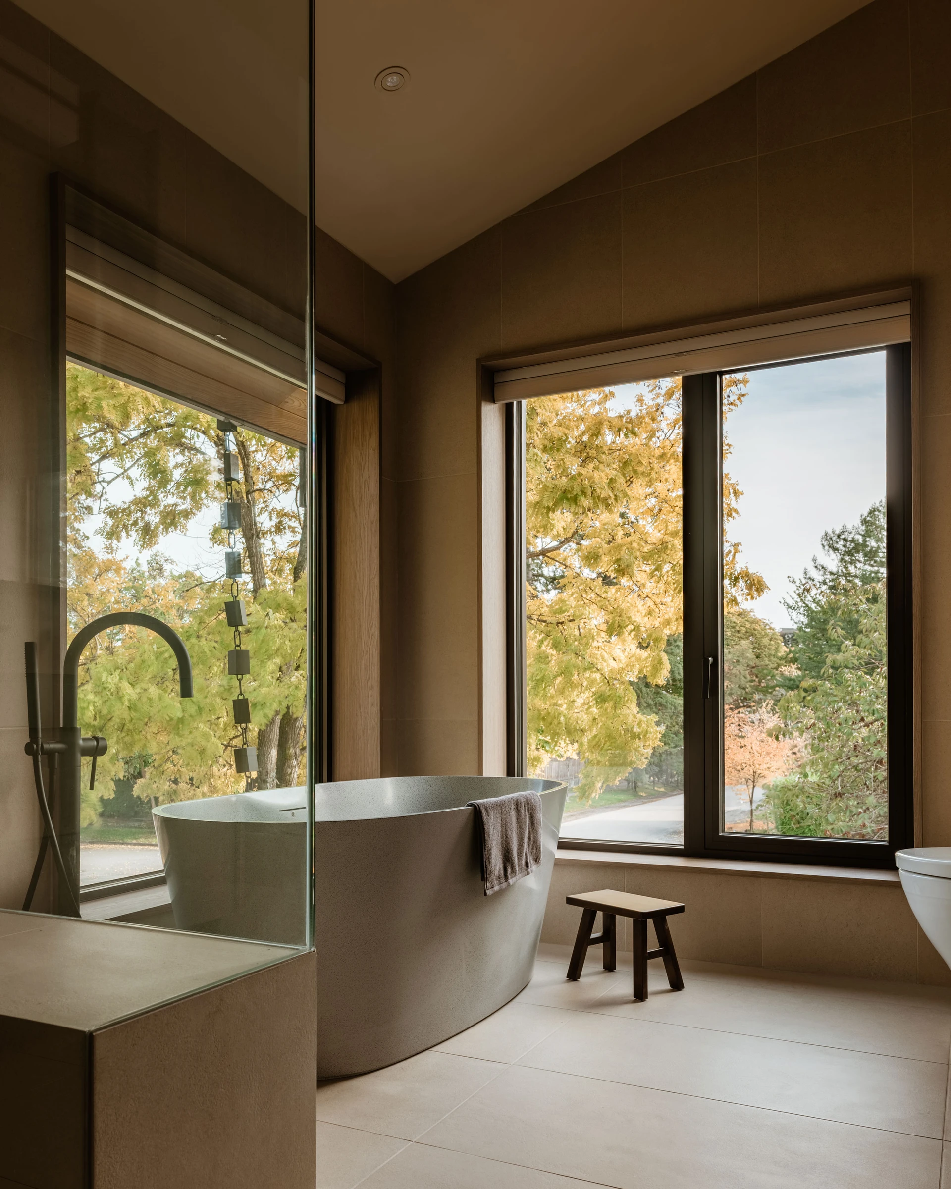 Modern bathroom with a freestanding bathtub, towel, small wooden stool, and large window showing autumn trees outside.