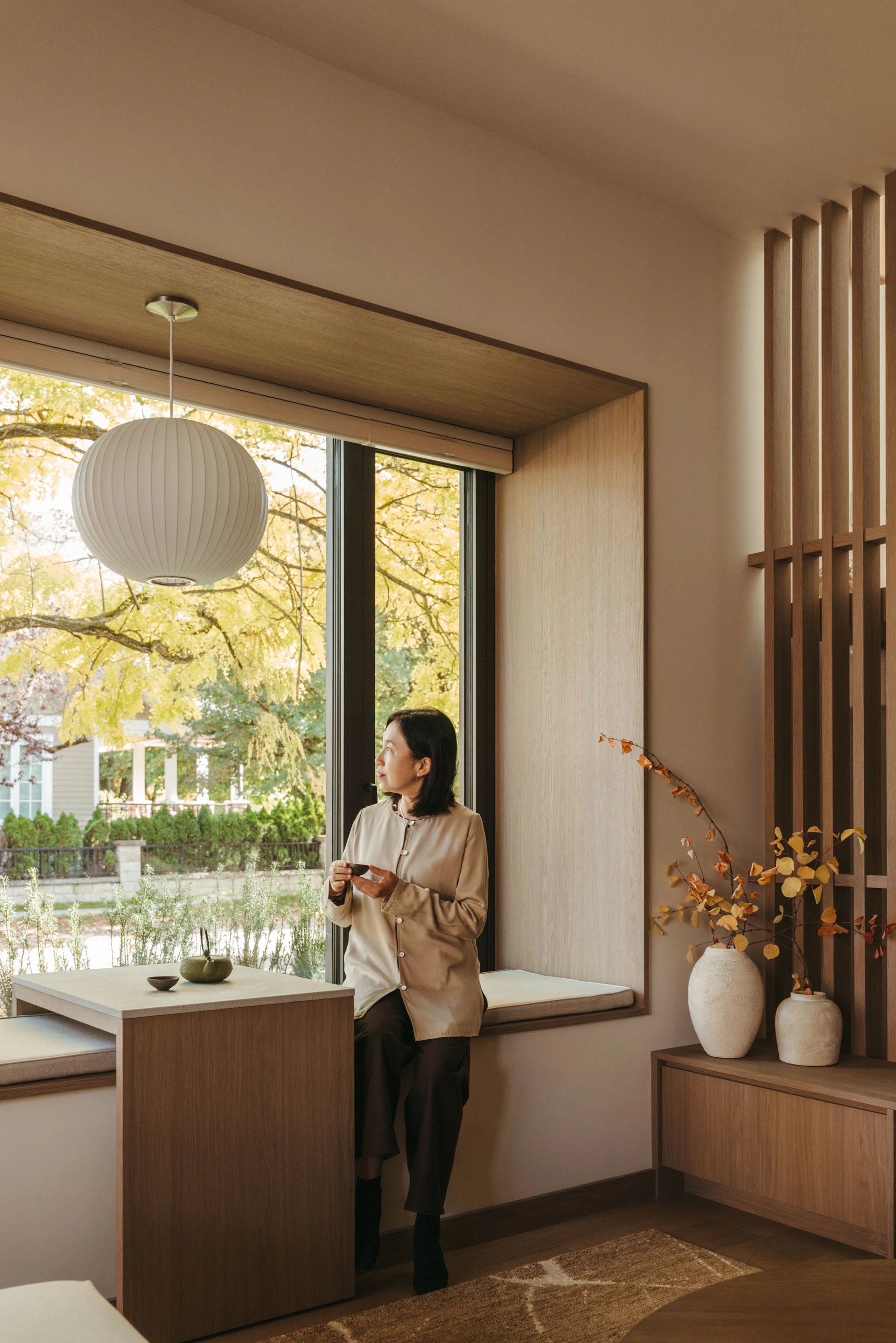 Woman sitting on a cushioned window seat drinking tea, looking out through large windows framed by autumn trees.