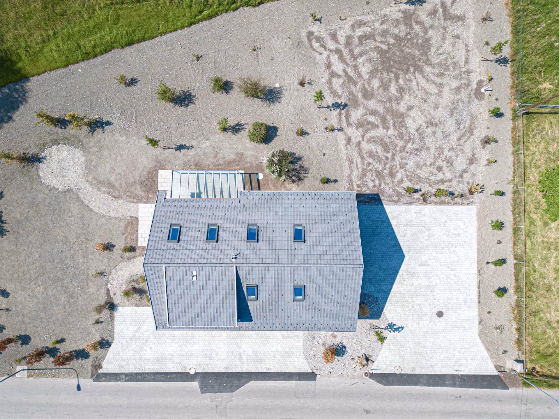Luftaufnahme eines modernen Hauses mit grauem Dach, umgeben von kargem Garten mit vereinzelten kleinen Büschen und gepflasterter Einfahrt.