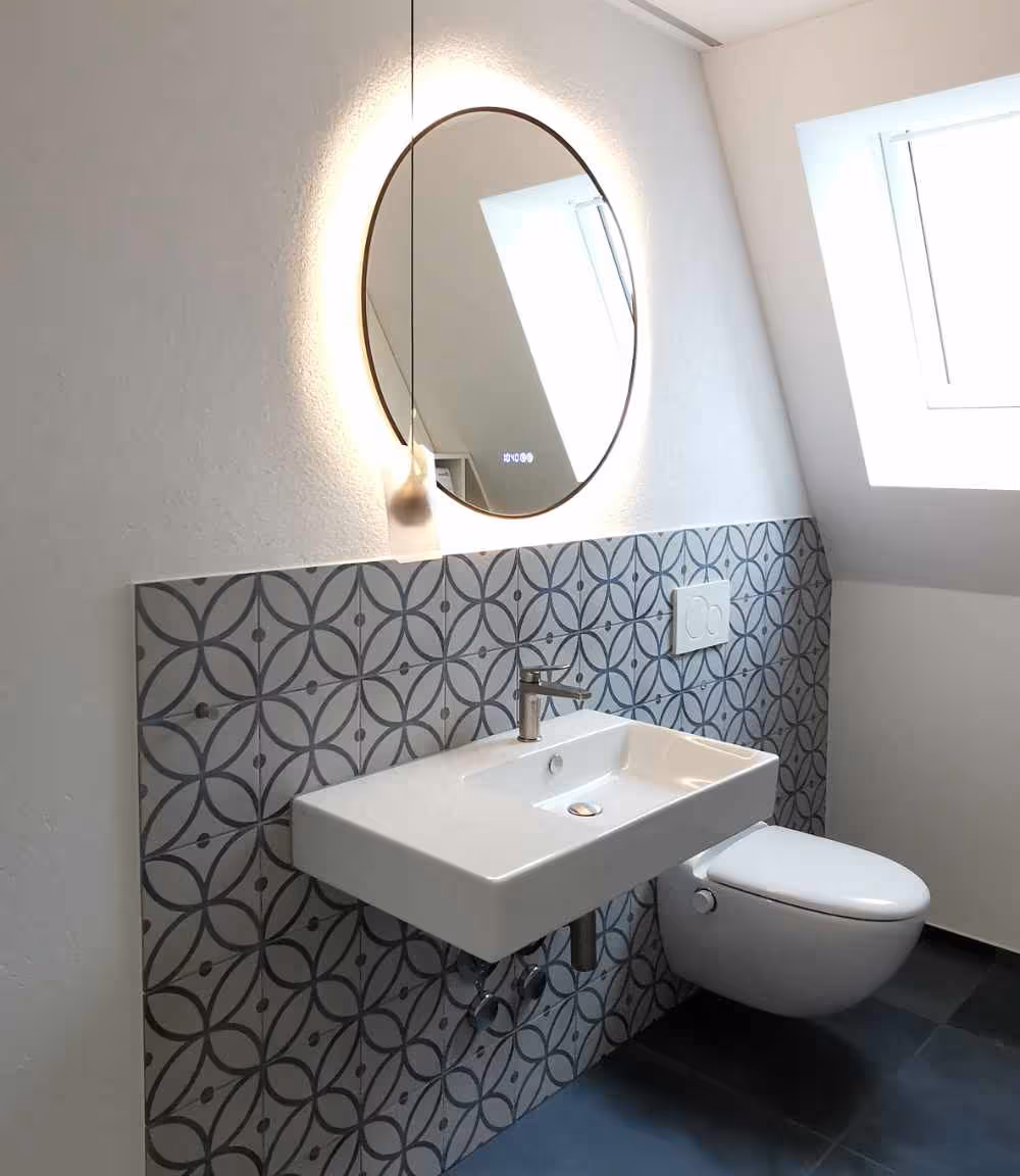 Modernes Badezimmer mit wandmontiertem rechteckigem Waschbecken, rundem beleuchtetem Spiegel, gemusterten Wandfliesen und wandhängendem WC unter einem Dachfenster.