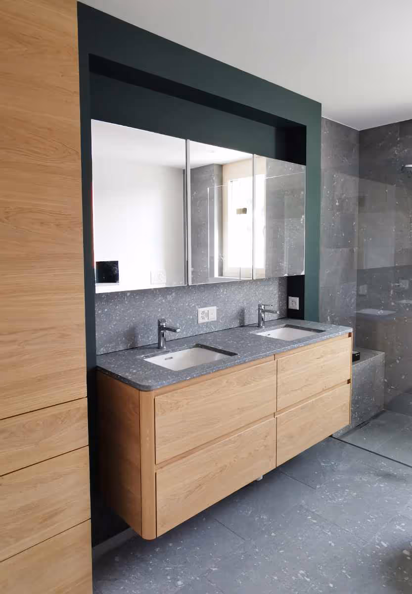 Modernes Badezimmer mit Doppelwaschbecken aus grauem Stein und hellen Holzschränken unter großen Spiegeln.