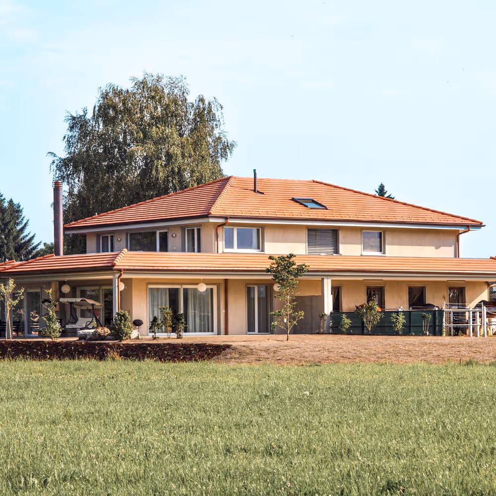 Modern zweistöckiges Haus mit rotem Ziegeldach und großer Veranda, umgeben von Rasen und Bäumen.