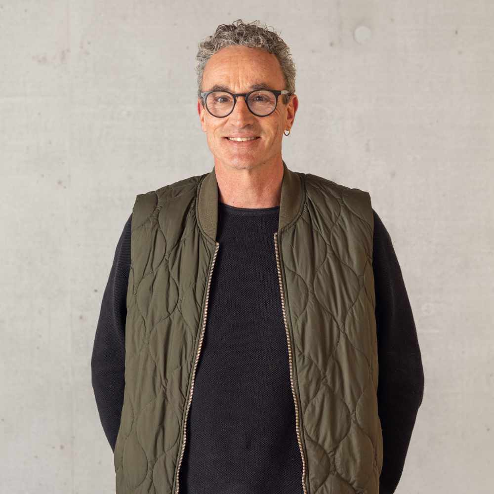 Mann mit kurzen grauen Haaren, Brille, Ohrring und grünem gesteppter Ärmellosem über schwarzem Pullover vor hellgrauer Wand.