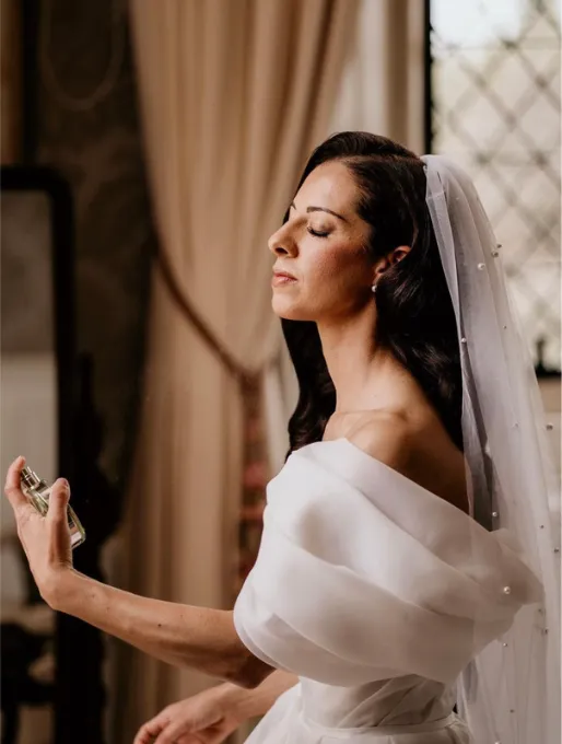 A beautiful bride, dressed in a white wedding gown, getting ready in our exquisite bridal suite at North Mymms Park. She is captured spraying perfume onto herself, adding a touch of elegance to her preparation. The suite's luxurious decor and soft lighting complement the bride's radiant smile, creating a charming and memorable moment as she prepares for her special day