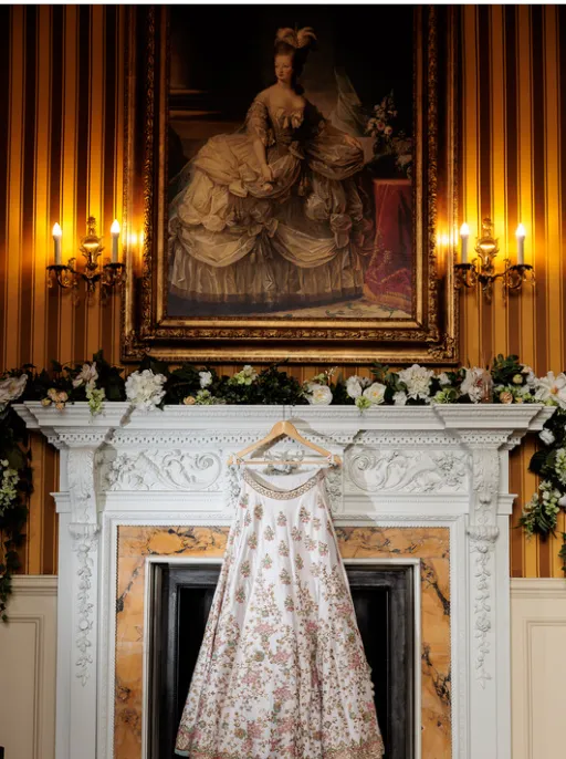 A traditional Indian wedding dress hanging on an ornate and grand fireplace inside the luxurious bridal suite at North Mymms Park. The fireplace is adorned with beautiful flowers, adding a touch of natural beauty and elegance to the opulent setting. Above the fireplace, a grand classic painting with a gold frame perfectly complements the gold and beige stripes of the wallpaper, further enhancing the luxurious ambiance of the room. The scene exudes timeless beauty and luxury, creating a charming and memorable setting for the bride's preparations
