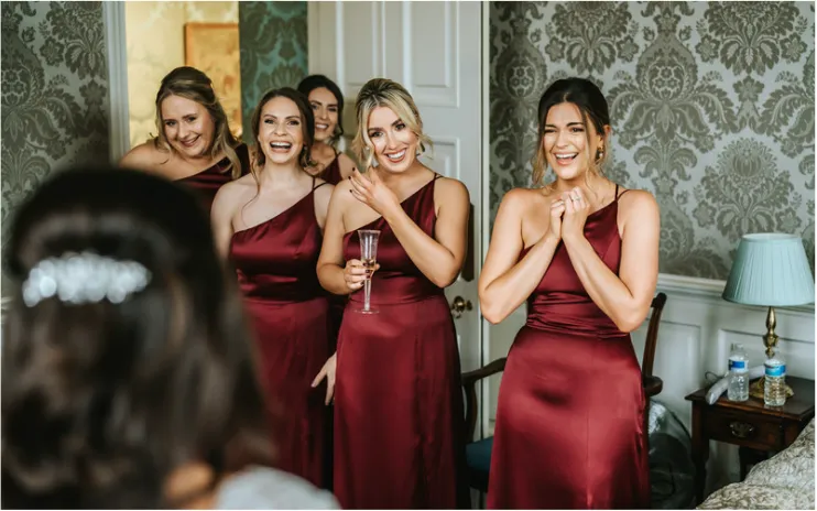 The bridesmaids seeing the bride for the first time, brimming with excitement and joy at North Mymms Park. Dressed in matching burgundy silk dresses, perfect for a Christmas wedding, their smiles light up the room. The luxurious setting of the bridal suite enhances the festive atmosphere, creating a heartwarming and memorable moment as they share in the bride's happiness on this special day