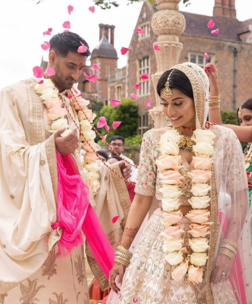 Joyful Asian bride and groom, surrounded by their beaming friends and family, celebrate their special moment at North Mymms Park's Mansion House grounds with a shower of vibrant Patel confetti