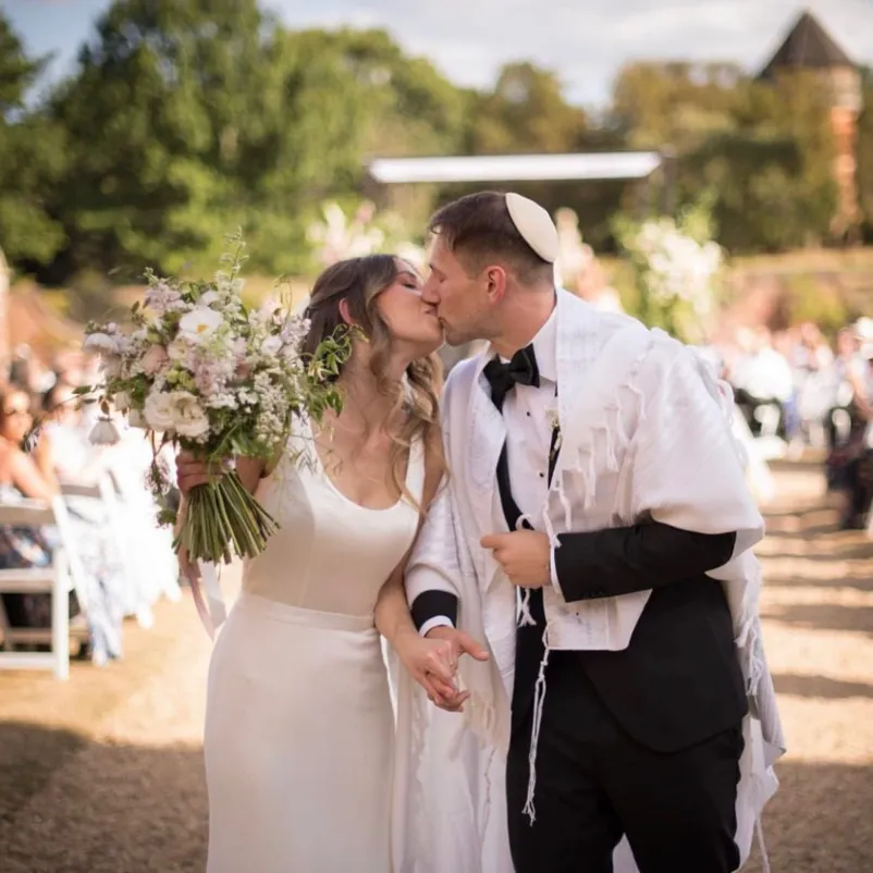 Embracing under their Chuppah, a joyful Jewish couple shares their first kiss after a heartwarming wedding ceremony in the manicured gardens of North Mymms Park, surrounded by the enchanting atmosphere of a summer wedding