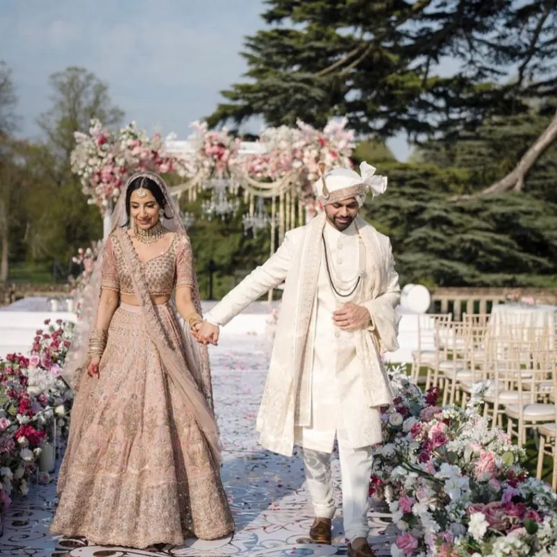 Hand in hand, a radiant Asian bride and groom share a moment of love after their religious ceremony in the stunning grounds of North Mymms Park. The beautiful mandap graces the background, adding to the charm of this summer wedding