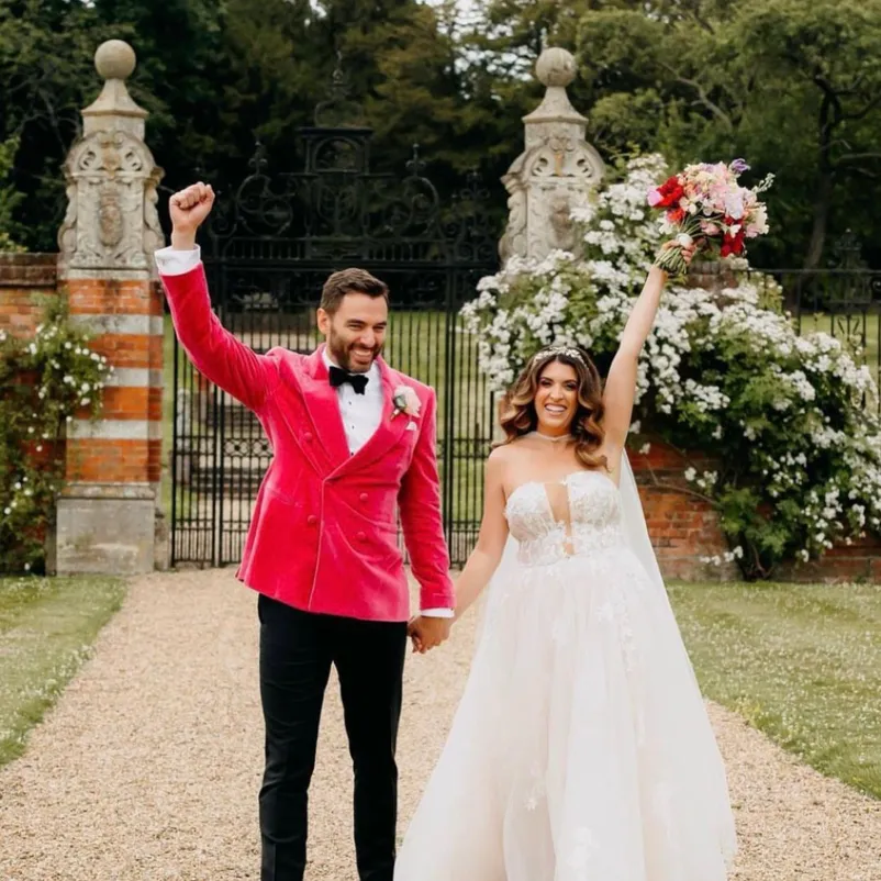 Hand in hand, a beaming bride and groom stand on the South Lawn of North Mymms Park, their other hands reaching towards the sky, smiling joyfully. The bride holds her wonderful bouquet aloft, adding to the enchantment, while a beautiful black gate adorned with roses forms a captivating backdrop to their delightful moment