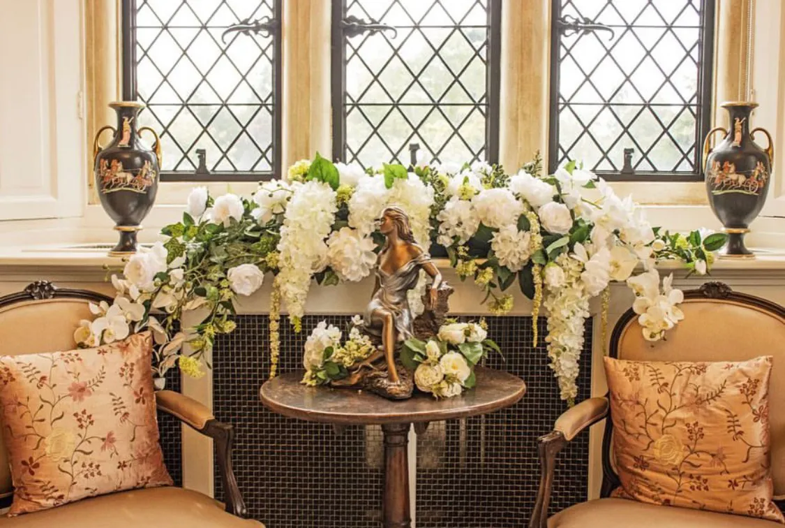 A close-up picture of the corner of the bridal suite at North Mymms Park. The image showcases a beautifully adorned table with an arrangement of flowers alongside a stylish statue of a woman adding a touch of artistry to the decor. The large windows in the background provide a view of the serene surroundings, inviting natural light into the room, creating a peaceful and elegant setting for the bride's preparations