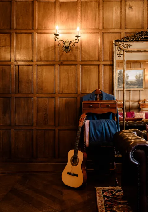 A close-up image of a corner in the oak room at North Mymms Park, which serves as the groom's room for getting ready. The oak-paneled walls exude a classic and refined atmosphere. Vintage ornate gold mirrors add a touch of elegance to the space. A stylish valet stands nearby, showcasing the groom's attire impeccably. A leather couch with a guitar completes the scene, providing a space for the groom to relax and perhaps strum a tune before his big day