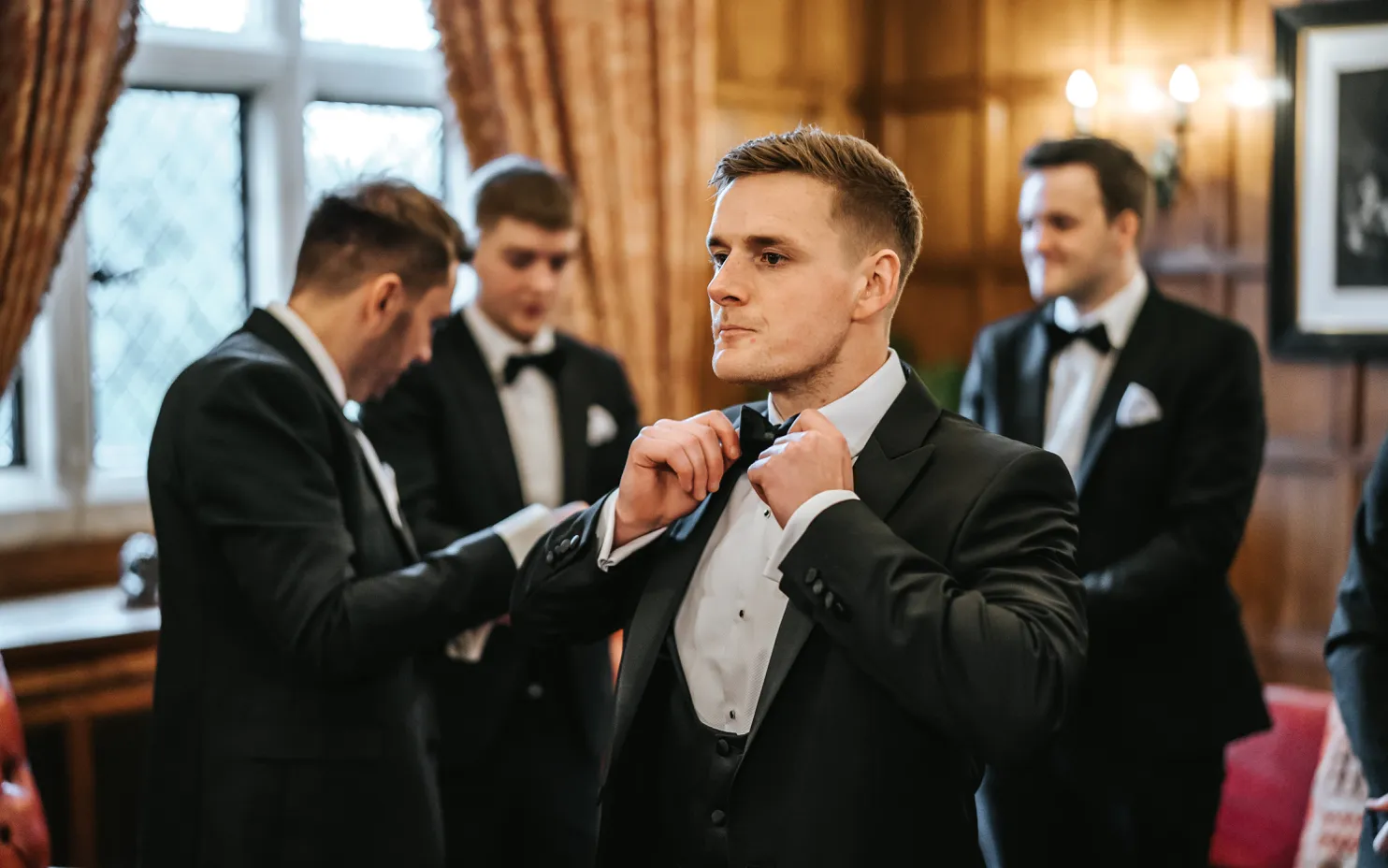 The groom, surrounded by his groomsmen, is fixing his bow tie in the luxurious oak room at North Mymms Park. The rich oak-paneled walls and ornate decor enhance the room's sophisticated ambiance. The camaraderie among the groom and his friends is evident as they prepare for the wedding, creating a warm and joyful atmosphere in this exquisite setting