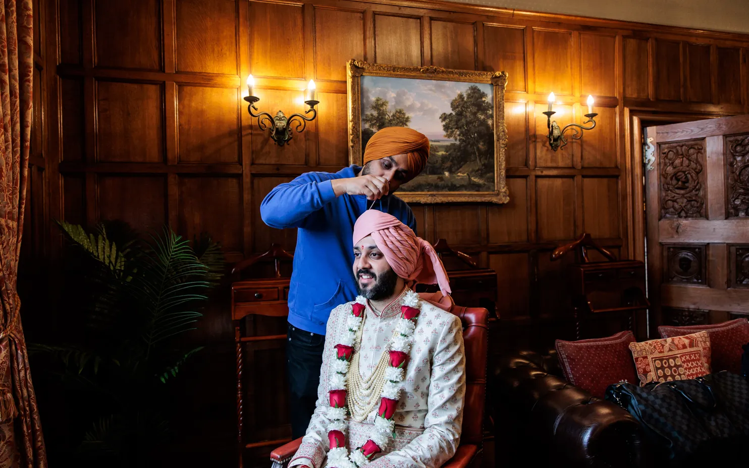 An Asian groom gets ready in the Oak Room at North Mymms Park for his religious ceremony. His best man is assisting him in fixing his turban, creating a moment of camaraderie and support before the auspicious event. The luxurious oak-paneled walls and elegant decor of the room add to the dignified atmosphere, creating a meaningful and memorable space for the groom's preparation for his special day