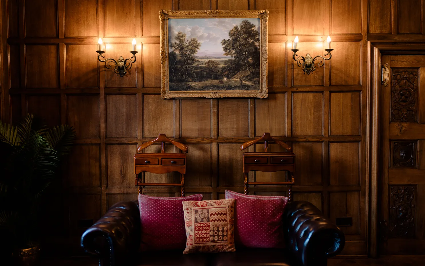A beautiful picture of the Oak Room at North Mymms Park. The room features oak-panelled walls that exude classic elegance. A big, classic painting adorns one of the walls, adding a touch of artistry to the space. Spot lighting accentuates the painting and creates a warm and inviting ambiance. Two valets stand nearby, ready to assist, and a brown leather couch with dark red cushions provides a comfortable and stylish seating area, making the Oak Room a refined and luxurious setting for any occasion