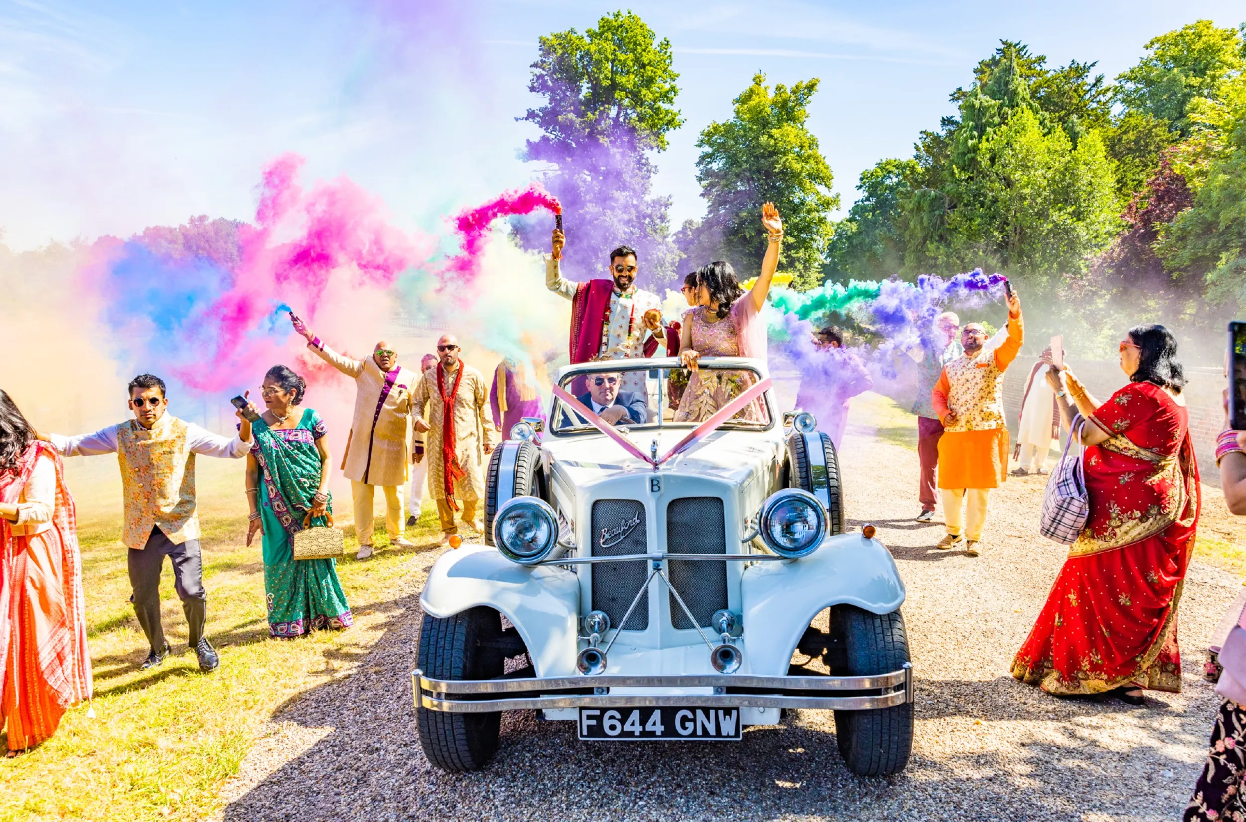 The groom makes a grand entrance on a classic car, surrounded by jubilant friends and family, to join the colorful Indian wedding festivities in the stunning grounds of North Mymms Park