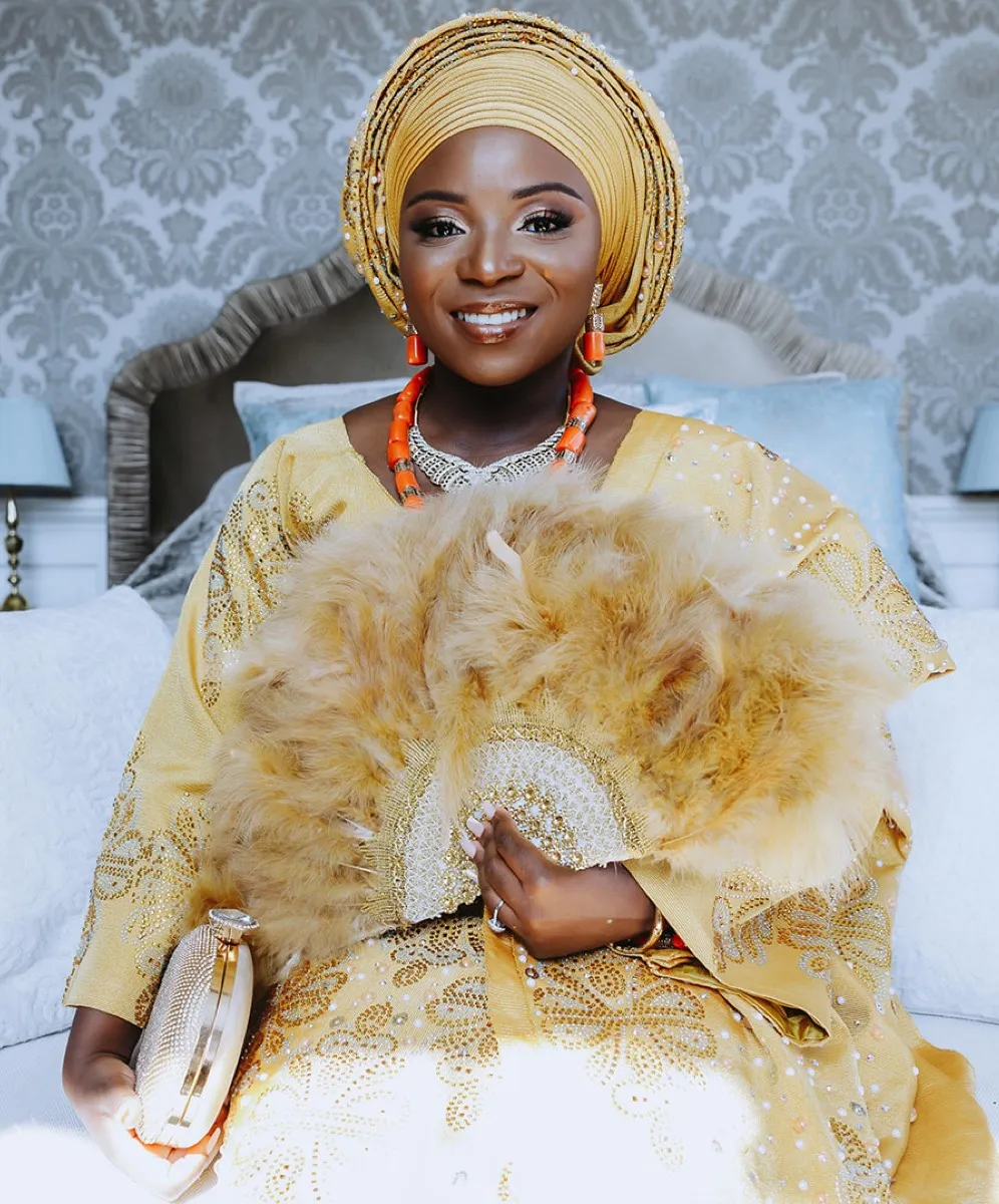 Beaming African Nigerian bride, adorned in a radiant yellow traditional wedding dress, holds a stylish clutch and a beautiful hand fan in her hand, captured in our stunning bridal suite at North Mymms Park
