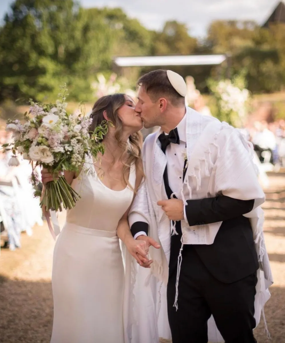 Embracing under their Chuppah, a joyful Jewish couple shares their first kiss after a heartwarming wedding ceremony in the manicured gardens of North Mymms Park, surrounded by the enchanting atmosphere of a summer wedding