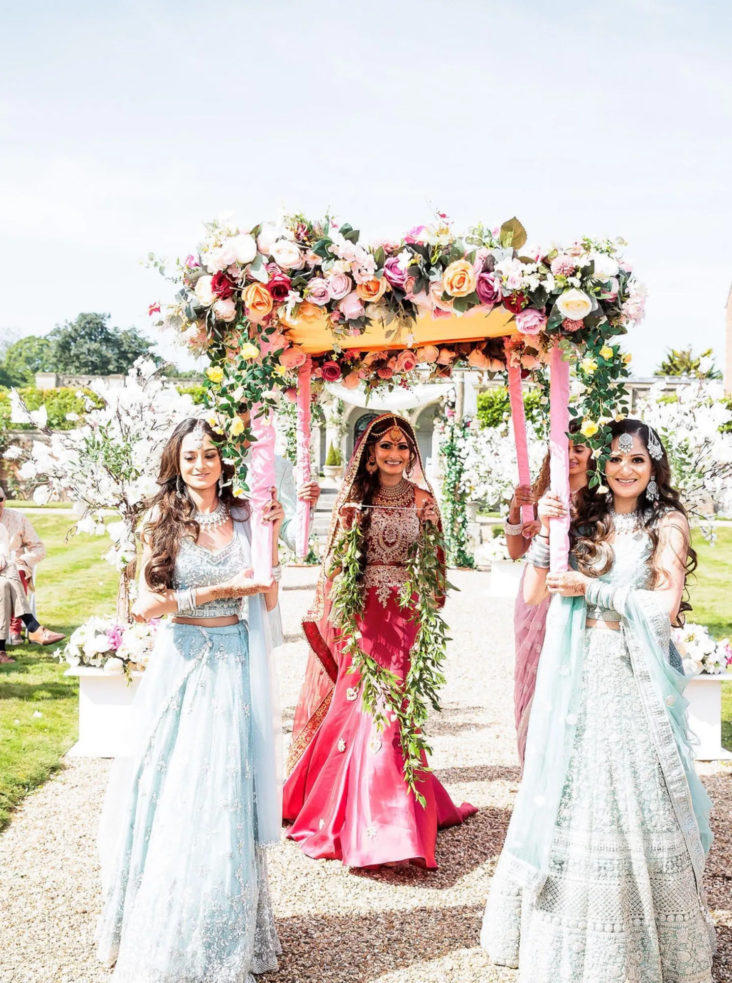 Joyful Pakistani bride, adorned in her traditional outfit, makes her way to her Nikkah ceremony under the floral canopy, surrounded by her friends and family, in the scenic gardens of North Mymms Park
