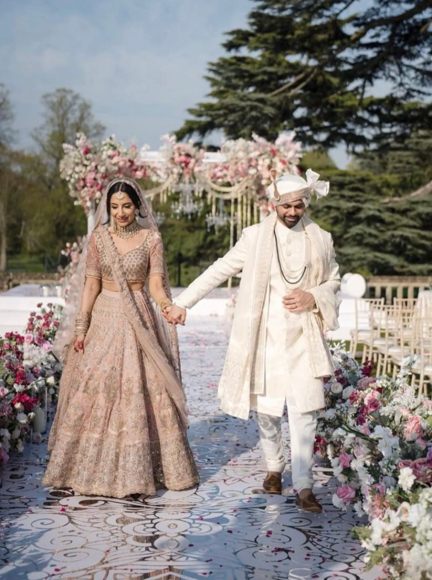 Hand in hand, a radiant Asian bride and groom share a moment of love after their religious ceremony in the stunning grounds of North Mymms Park. The beautiful mandap graces the background, adding to the charm of this summer wedding