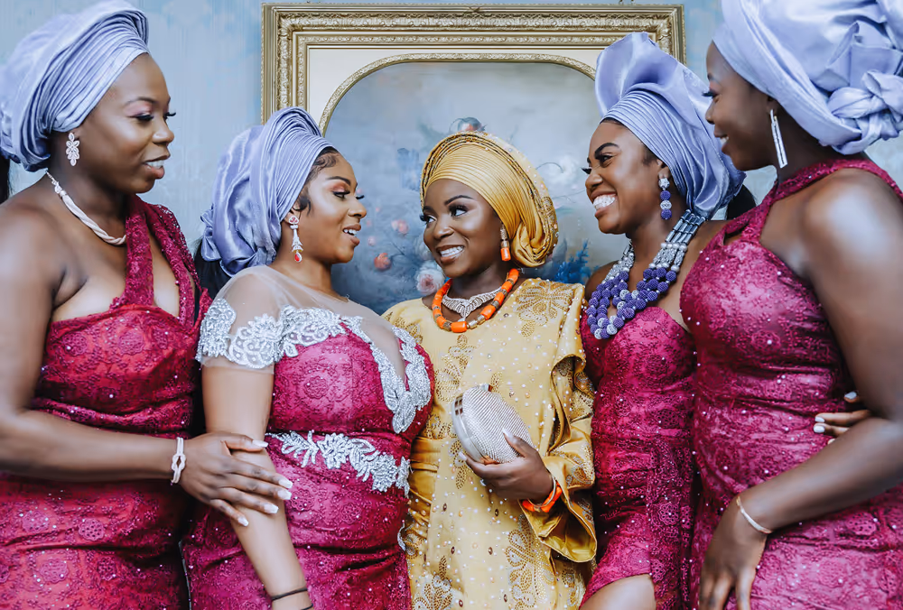 Radiant African bride in a vibrant yellow traditional bridal outfit, surrounded by her bridesmaids dressed in elegant purple Nigerian attire, captured in the picturesque Bridal suite at North Mymms Park