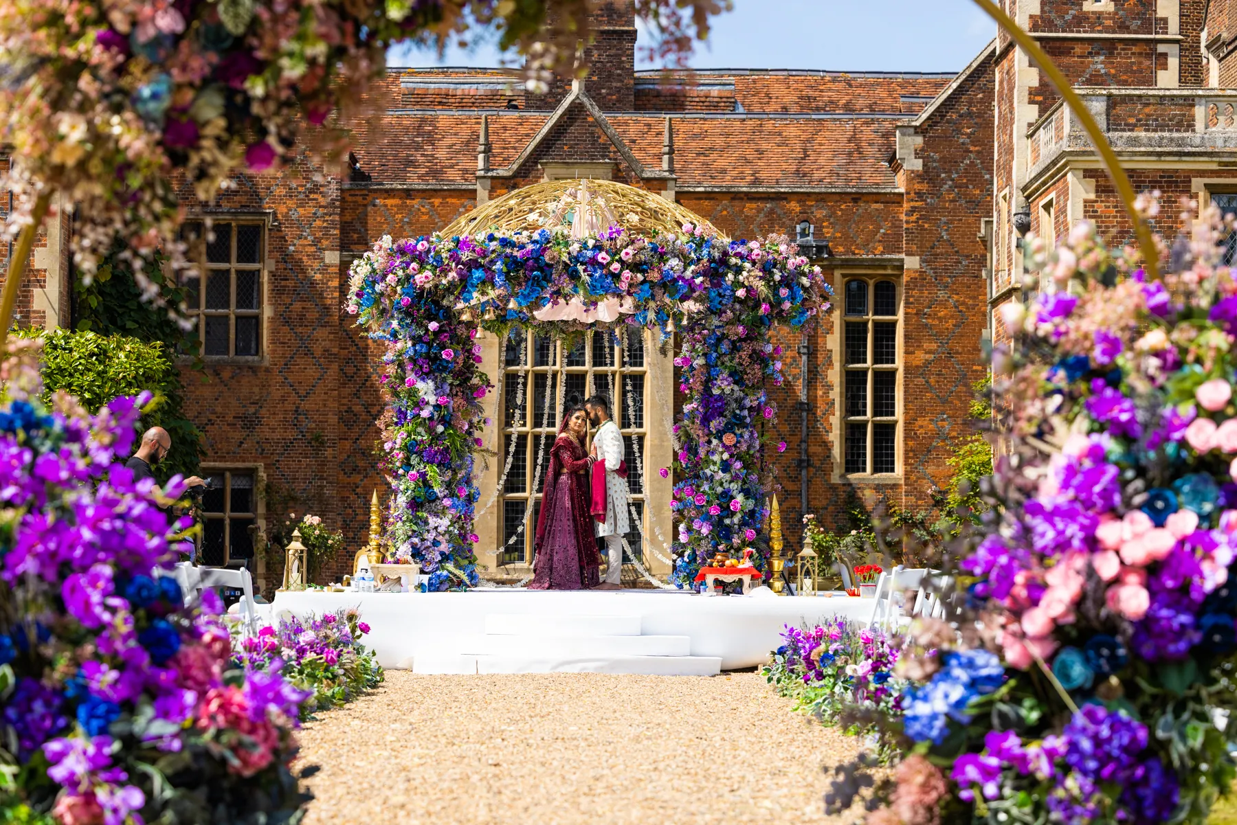 Captivating summer wedding at North Mymms Park, featuring an Asian couple in traditional attire, standing under a beautifully adorned mandap. The splendid mansion house in the background adds to the enchanting atmosphere of the celebration