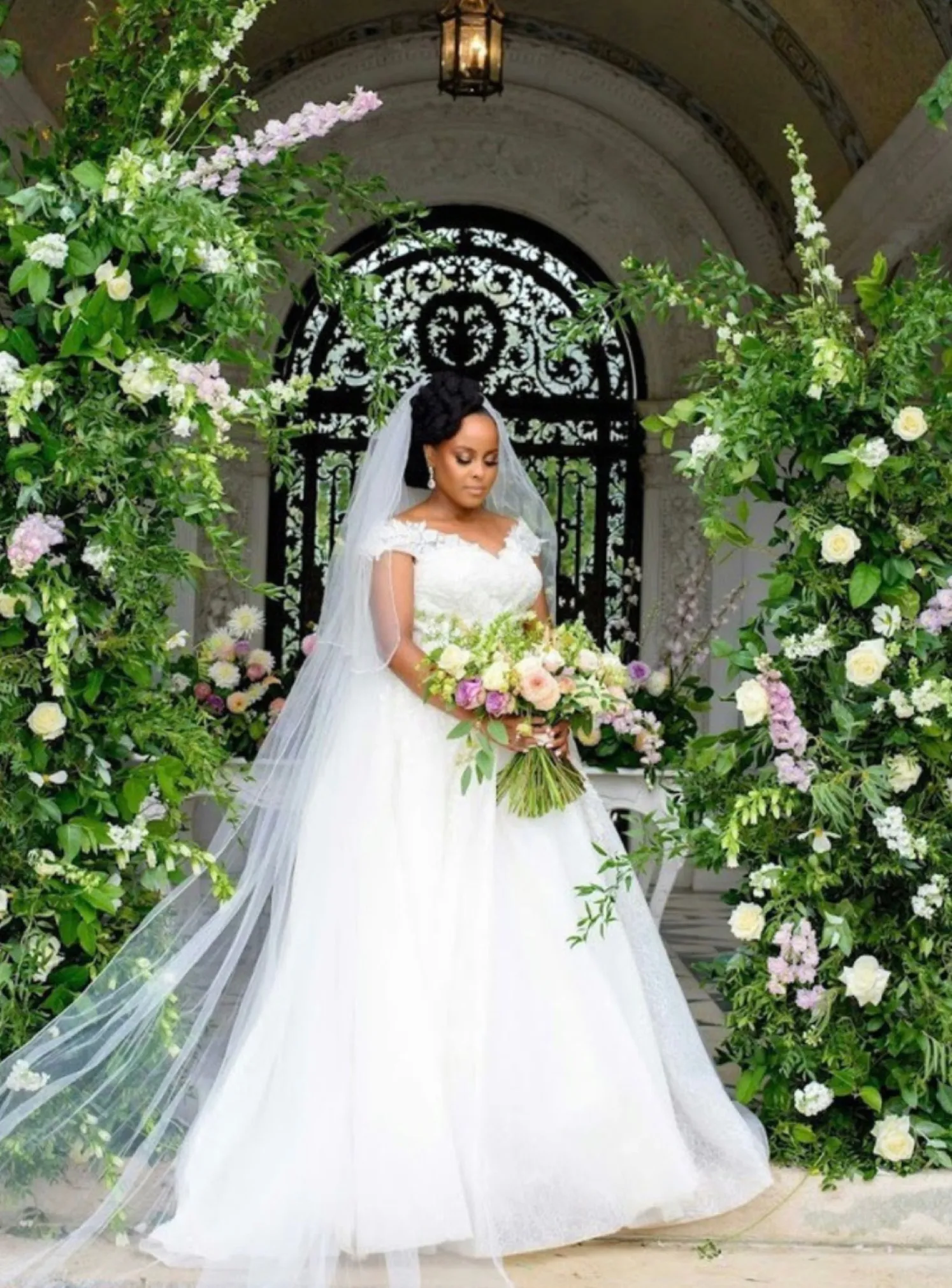 Surrounded by a captivating floral arch, a stunning African bride shines in the Loggia at North Mymms Park during a beautiful summer wedding celebration