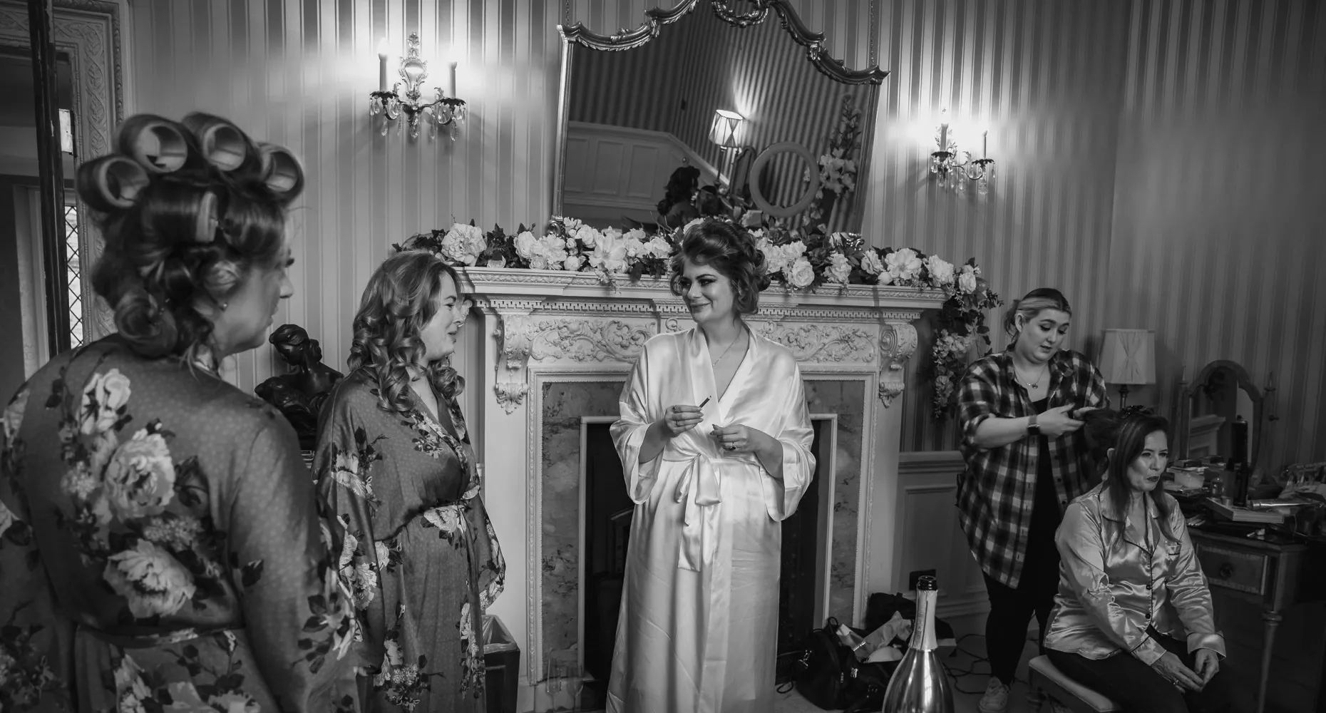 A black and white image capturing the bride and her bridesmaids getting ready in the bridesmaids' room at North Mymms Park. They are wearing matching silk robes, with their hair adorned with big, elegant hairpins. A hairstylist is seen working on one of the bridesmaids, adding to the excitement and joy of the moment. The happy and cheerful atmosphere reflects the anticipation and love shared by the bride and her closest friends on this special day
