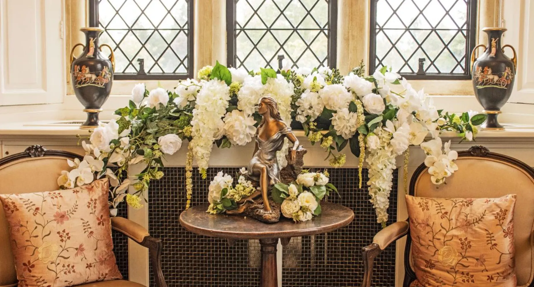 A close-up picture of the corner of the bridal suite at North Mymms Park. The image showcases a beautifully adorned table with an arrangement of flowers alongside a stylish statue of a woman adding a touch of artistry to the decor. The large windows in the background provide a view of the serene surroundings, inviting natural light into the room, creating a peaceful and elegant setting for the bride's preparations.
