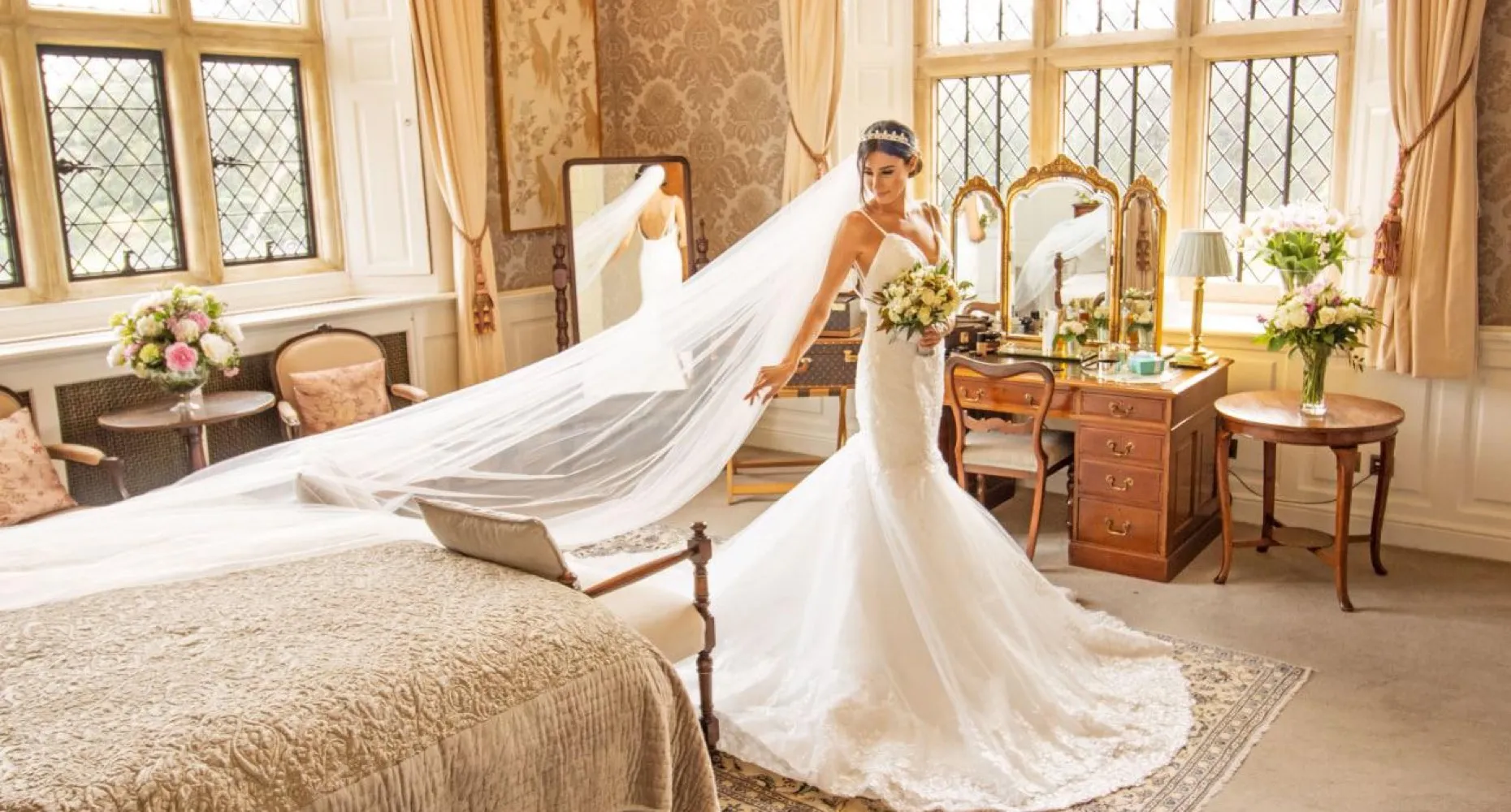 A stunning English bride getting ready in our exquisitely appointed bridal suite at North Mymms Park. She stands in front of a big mirror, dressed in an elegant white gown with a long veil cascading gracefully. The luxurious and tastefully decorated suite provides a beautiful backdrop as the bride prepares for her special day, radiating elegance and beauty