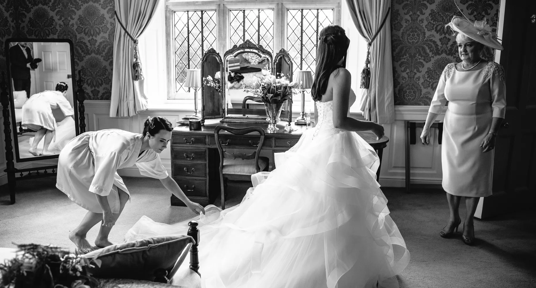 A black and white image of an elegant English bride in the stunning bridal suite at North Mymms Park, all ready for her special day. Her mother, dressed beautifully in a formal hat and dress, gazes fondly at her daughter. In the background, her sister is seen adjusting the bride's dress skirt with care. The monochrome tone adds a classic and timeless feel to the heartfelt and joyous moment before the wedding