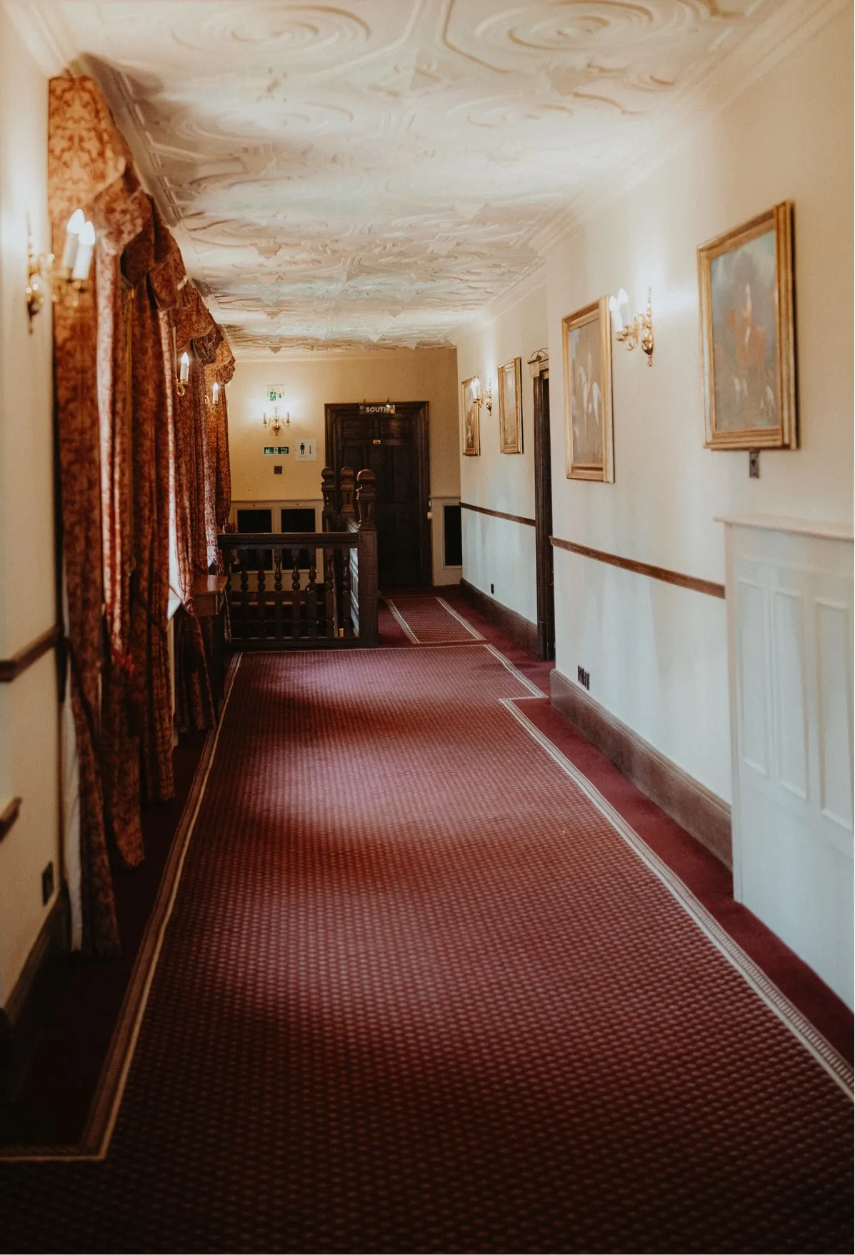 Grand hallway adorned with old paintings on its walls and a dark red carpet