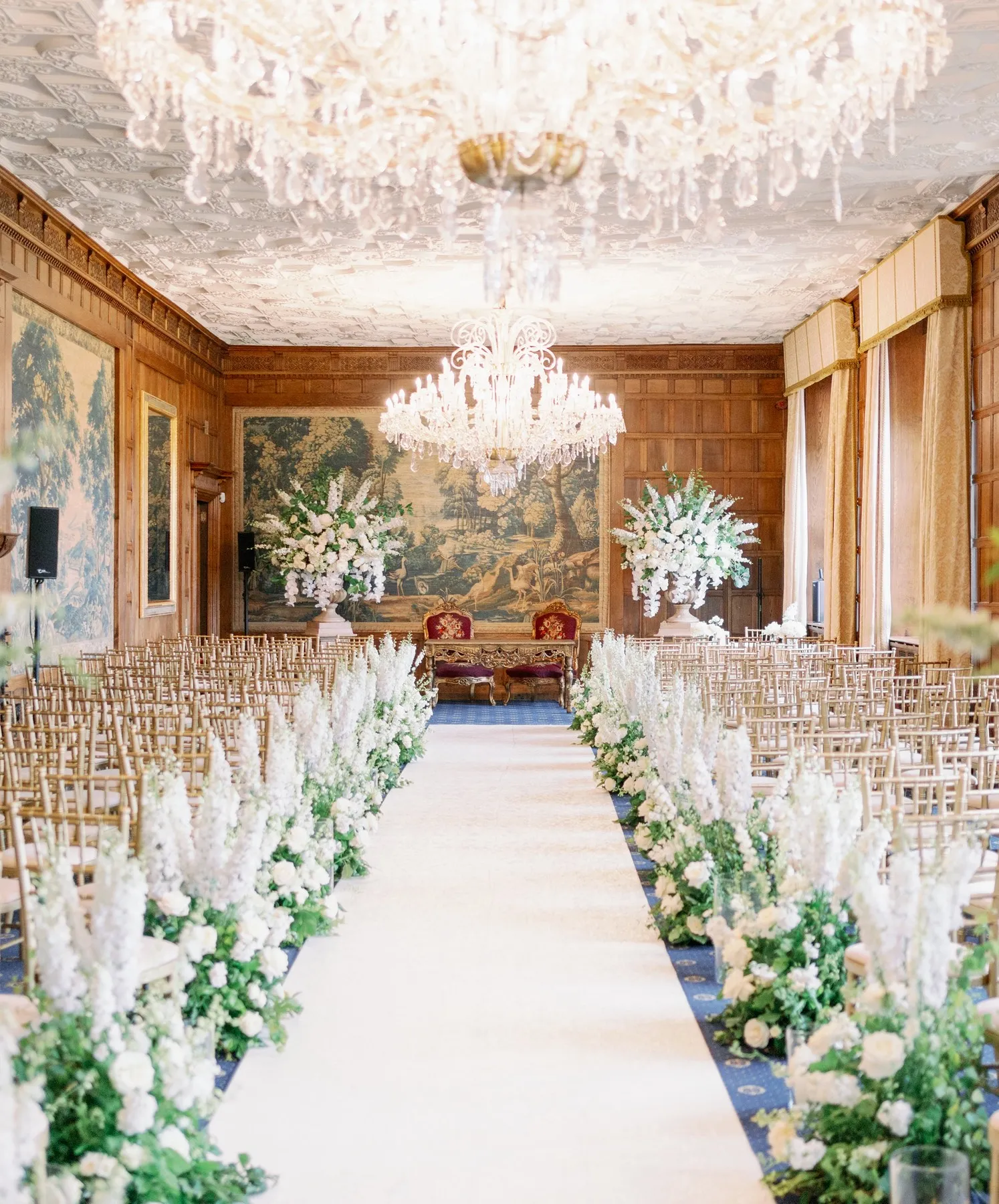 Romantic grand room in historical country house with big sparkling chandeliers, oak panels, and tapestries, set up for a civil ceremony with lavish floral decor
