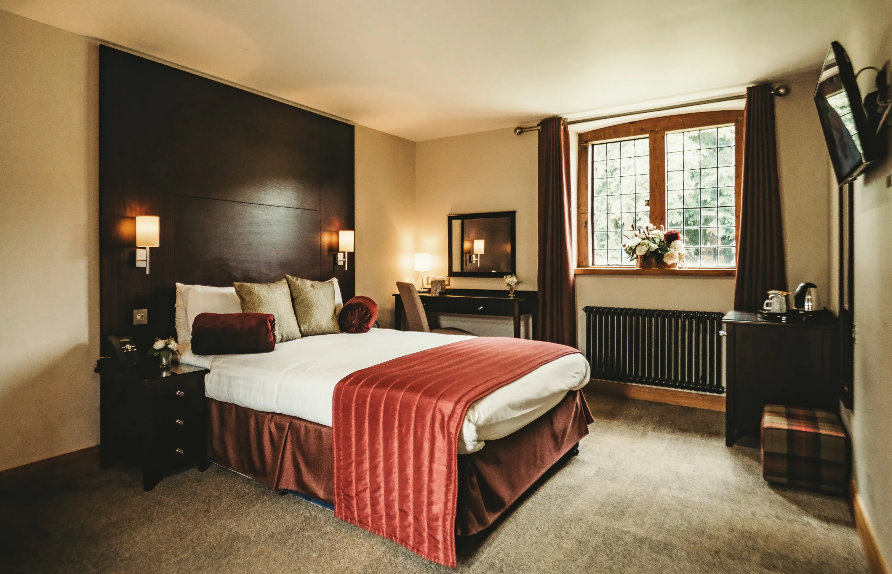 A modern guest bedroom in the stables at North Mymms Park. The room is equipped with a comfortable double bed with cushions, a sleek TV, a convenient dressing table, and coffee facilities. Large mirrors adorn the room, and an en-suite bathroom completes the inviting setup, providing a perfect space for guests to relax and unwind